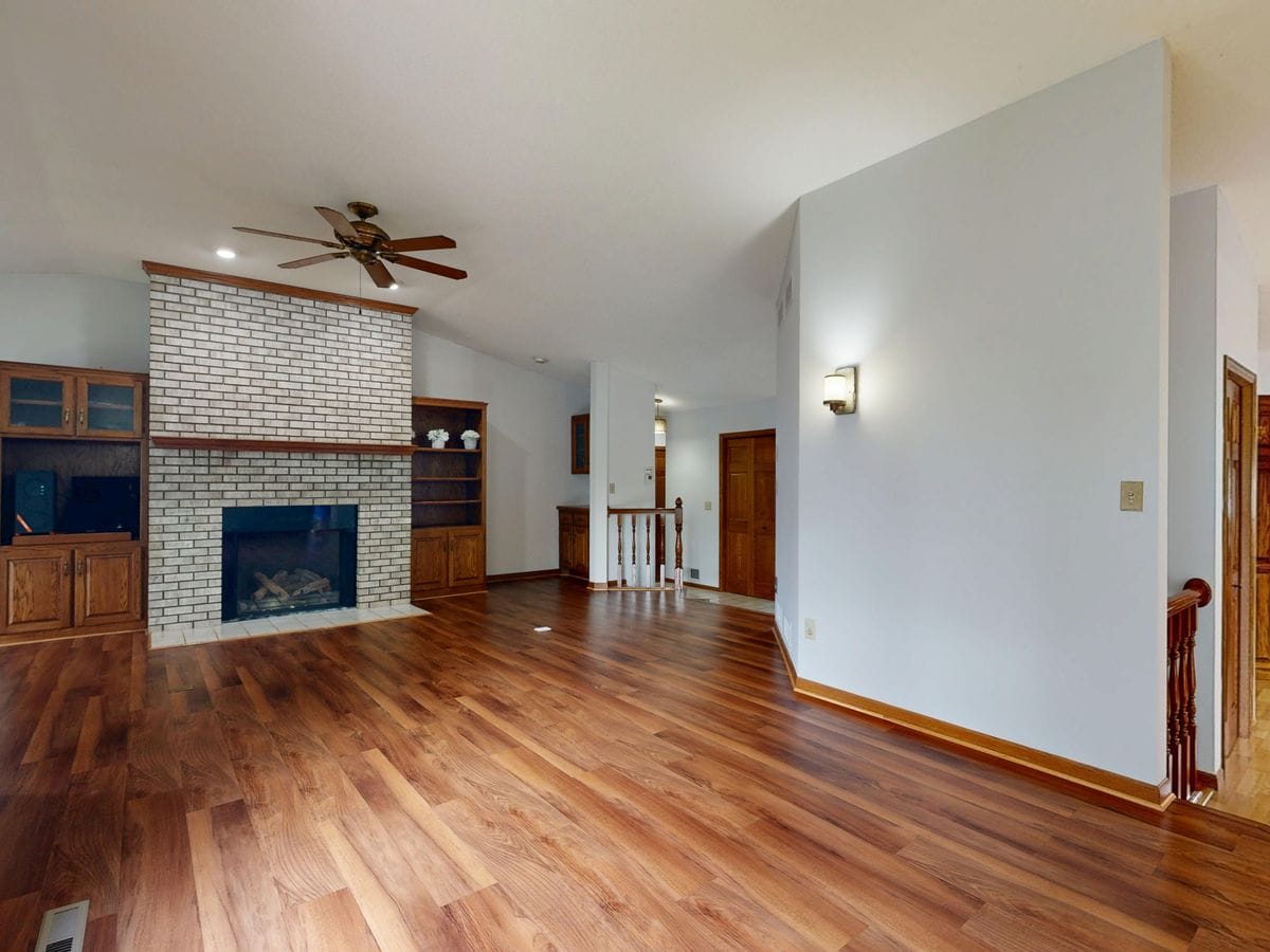 An empty living room with a painted brick fireplace, built-in shelving, and wood floors