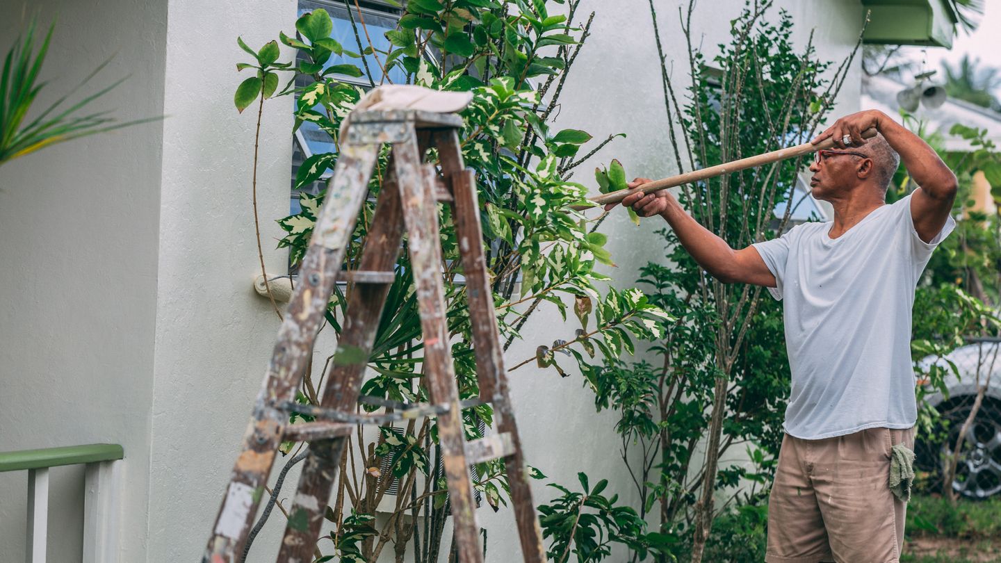 A painter rolling paint onto the exterior wall of a house beside a ladder